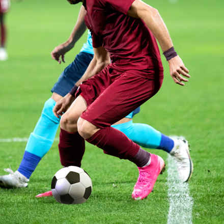 Picture of live football game, two pairs of legs from opposing teams tackling a football.