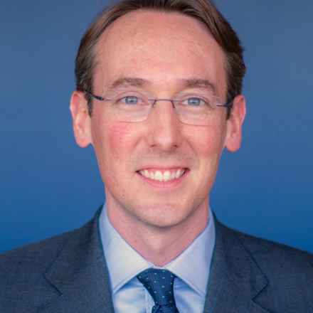 Headshot of Dr Keenan, a white man smiling at the camera wearing glasses and shirt, tie and suit. he is sitting against a blue background