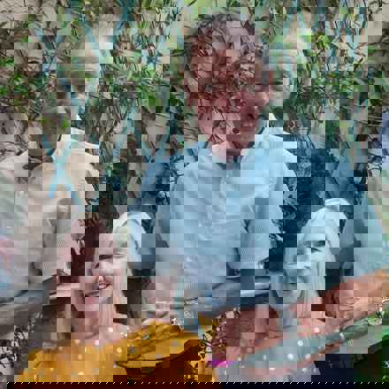 Peter and wife sue posing to camera. Peter standing smiling wearing a light blue shirt, with his arms around wife sue who is sat down wearing yellow spotted dress